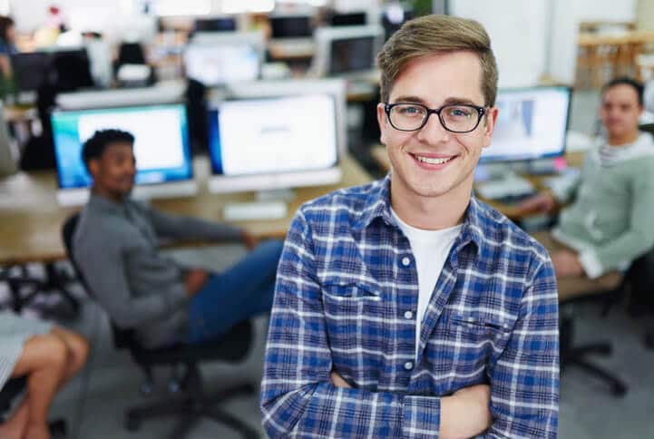 young man in a blue shirt smiles with computers behind him