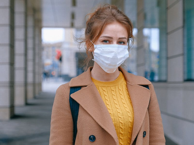 a woman in a tan jacket and yellow sweater wears a face mask