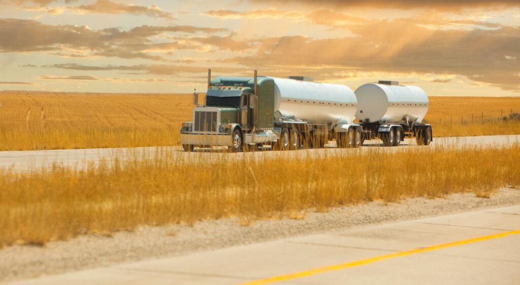 Semi-truck hauling fuel down a highway through wheat fields.